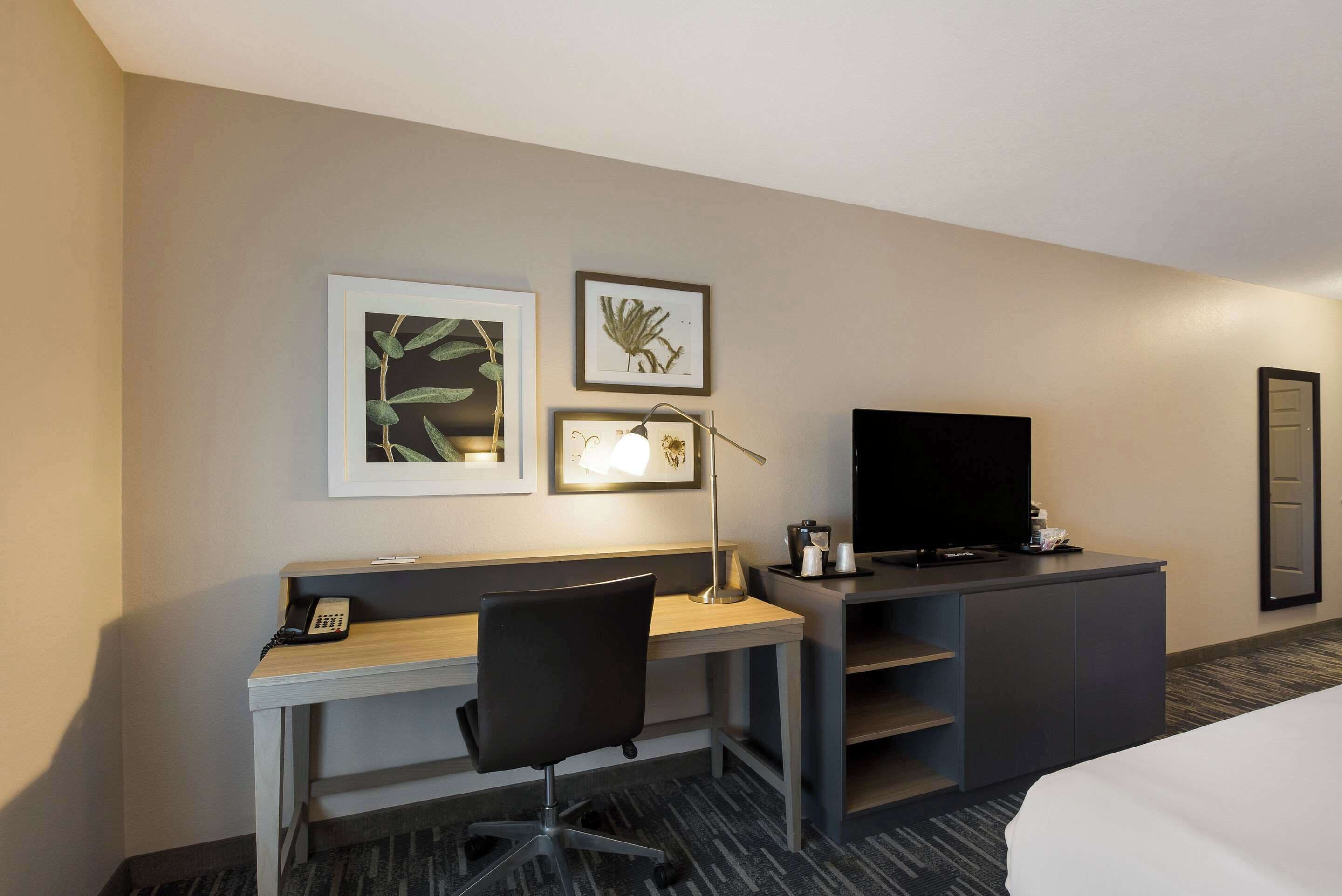 A hotel room with a desk, chair, television, and artwork on the wall.