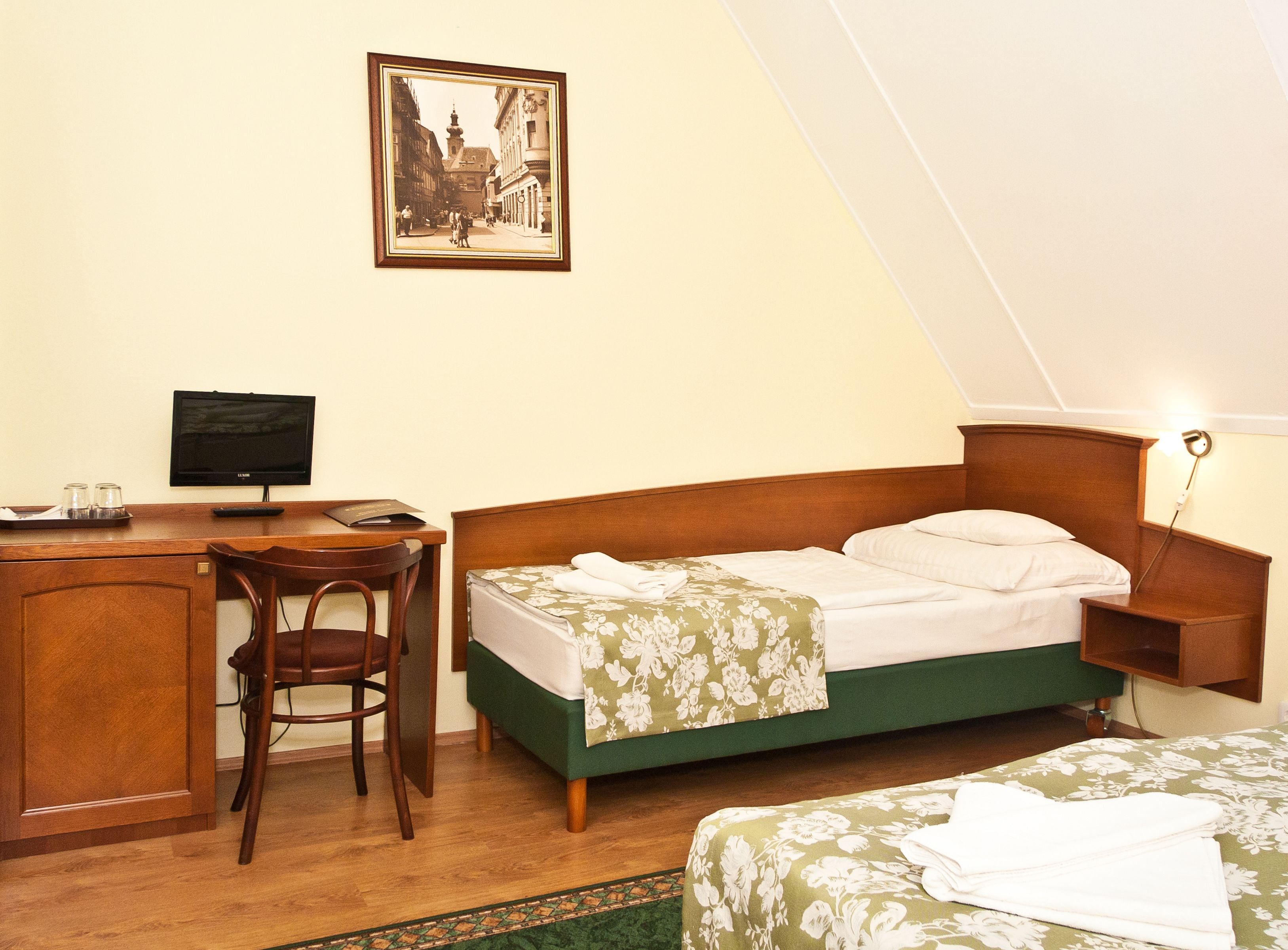 Quarto de hotel com escrivaninha de madeira, uma cadeira, uma cama com colcha estampada de flores verdes e um quadro na parede.