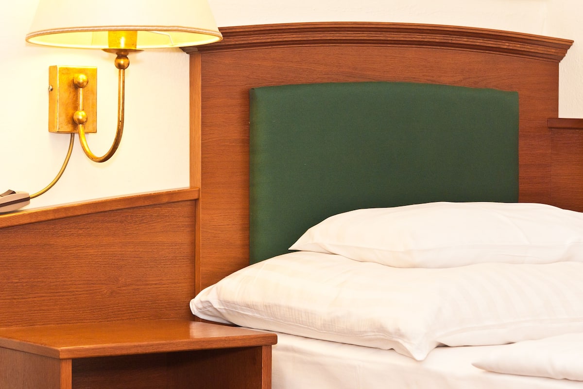 Una cama bien hecha con sábanas blancas, un cabecero tapizado en verde y una mesita de noche con una lámpara.