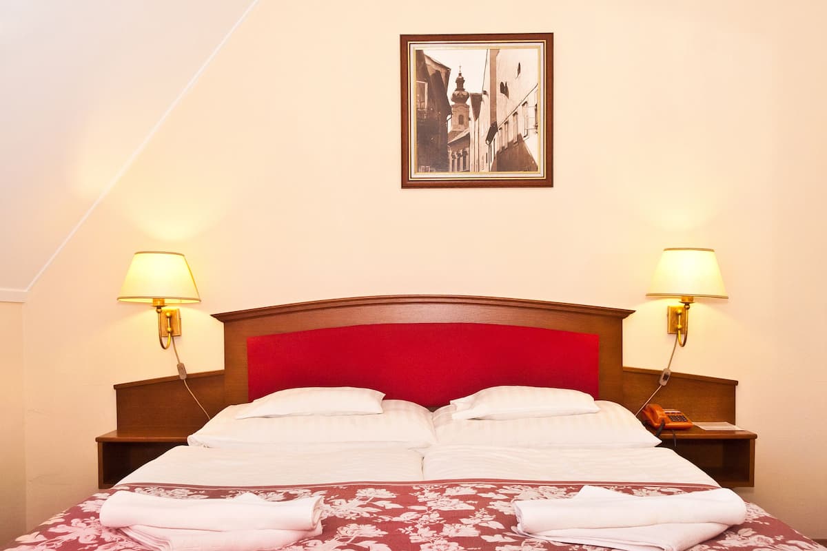 Habitación de hotel con cabecera roja, dos lámparas de noche, un cuadro en la pared y camas ordenadas con toallas.