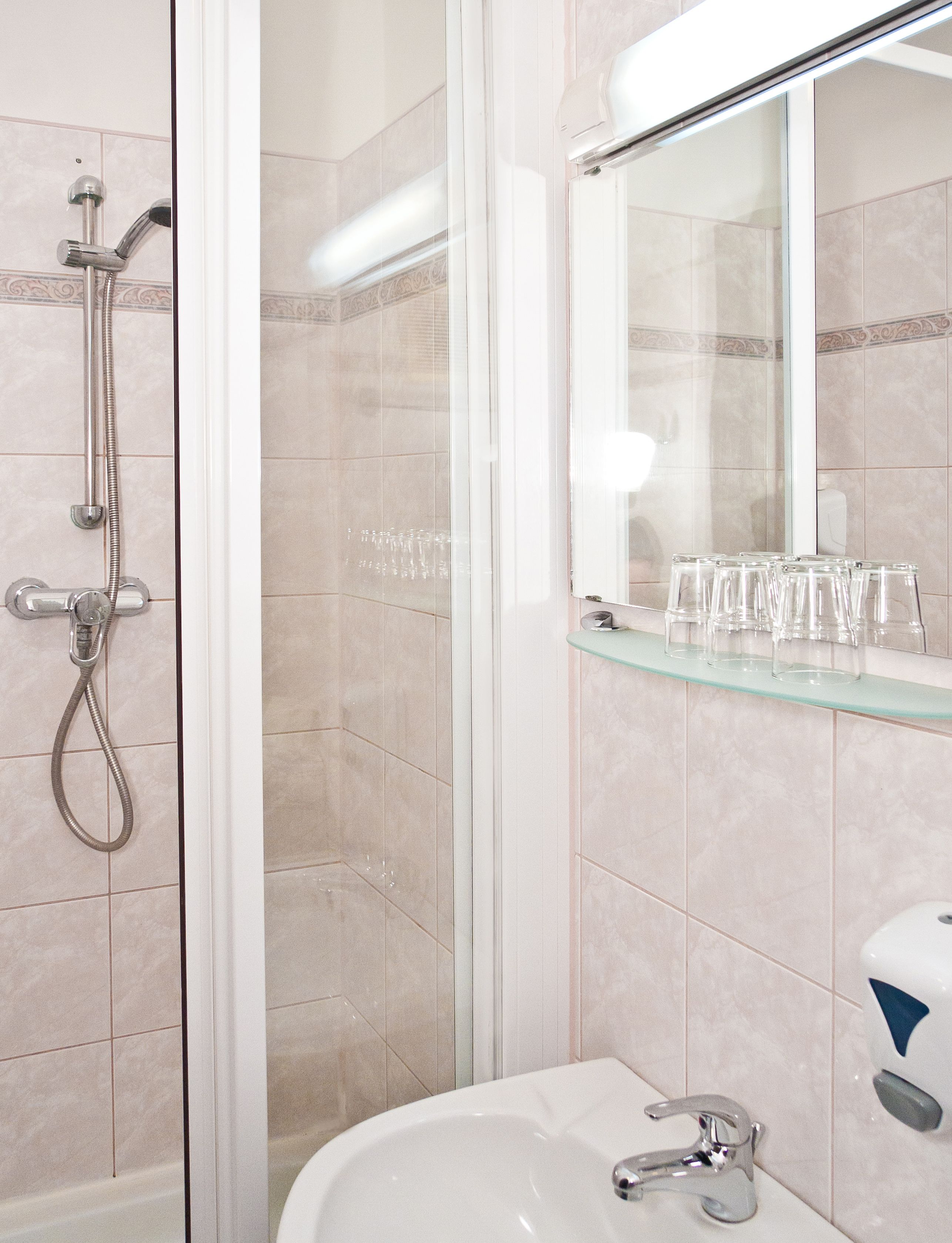 Baño con ducha, lavamanos y repisa de vidrio con vasos.