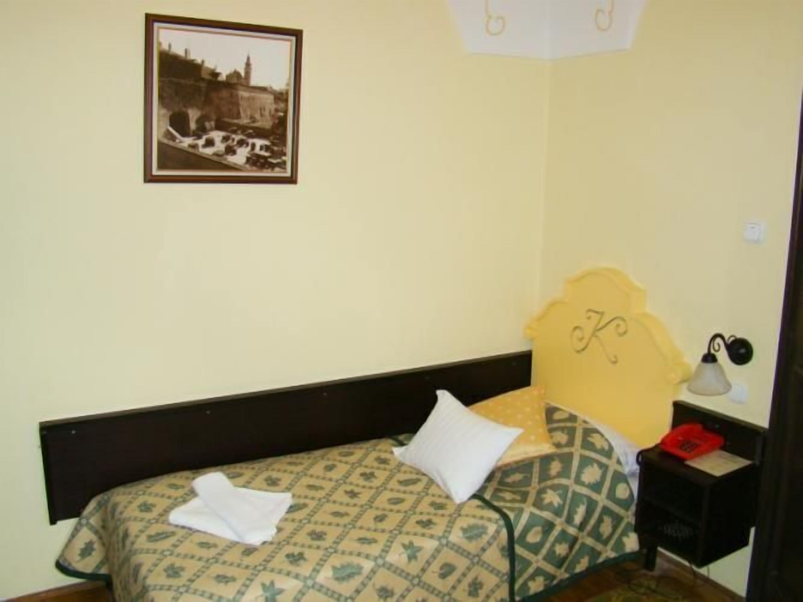 Ein kleiner, schlichter Raum mit einem Bett, einem Nachttisch, einer Lampe und einem gerahmten Bild an der Wand.