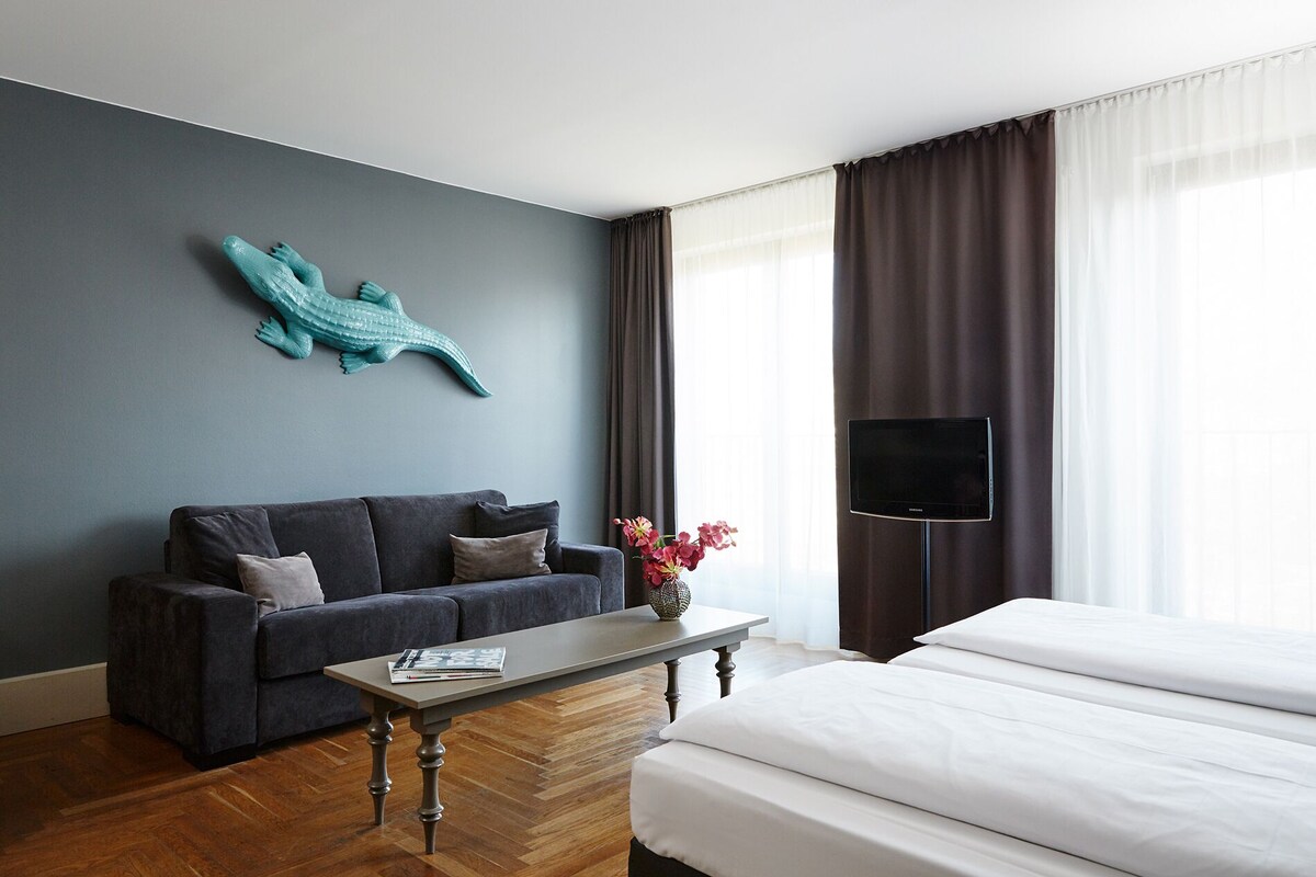 Une chambre d’hôtel avec un mur bleu foncé, une décoration représentant un crocodile turquoise, un canapé gris, un lit blanc, une table basse en bois, un vase avec des fleurs, un téléviseur à écran plat et une fenêtre avec des rideaux.