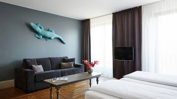 Habitación de hotel con una pared azul oscuro, un adorno de cocodrilo color turquesa, un sofá gris, una cama blanca, una mesa de centro de madera, un florero con flores, un televisor de pantalla plana y una ventana con cortinas.