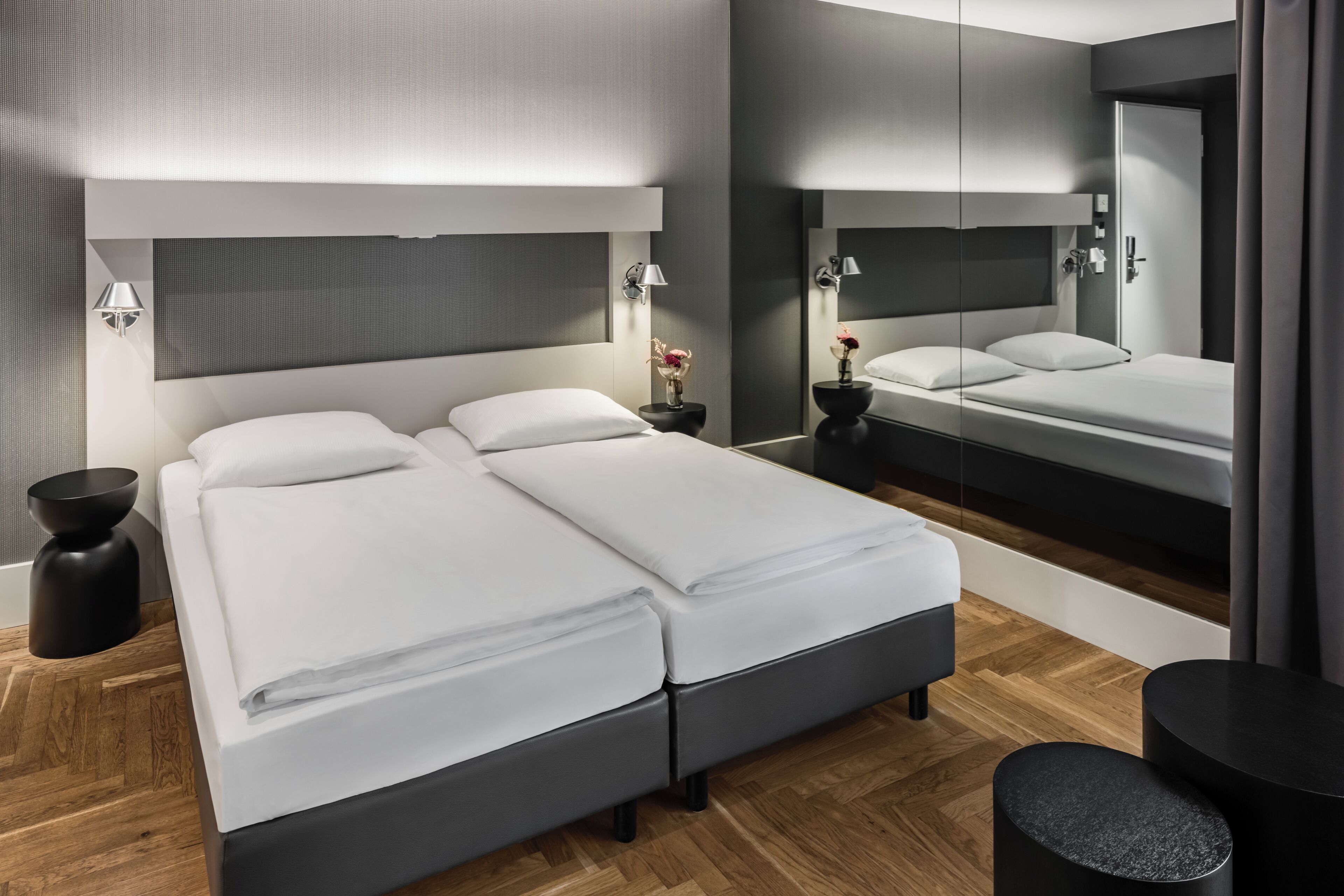 A modern hotel room with a large bed, bedside tables, a mirror, and a wooden floor.