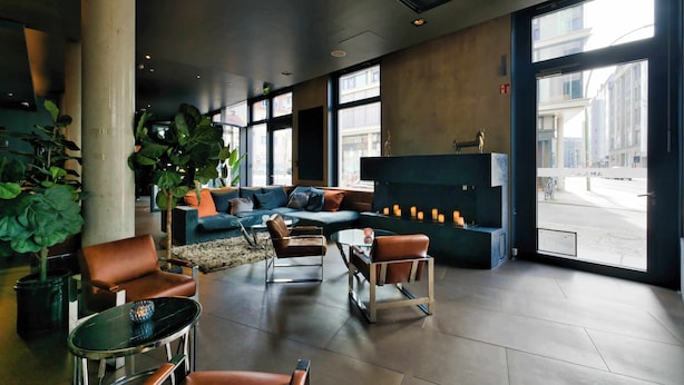 A modern lobby with a sofa, chairs, a coffee table, large windows, and a fireplace. - Hotel AMANO Rooms & Apartments (Berlin)
