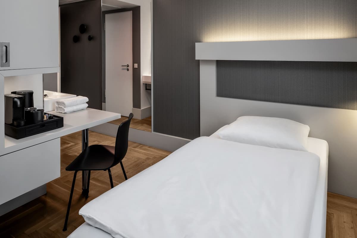 Una habitación de hotel moderna con una cama individual, un escritorio con silla, cafetera y una mesita con toallas.