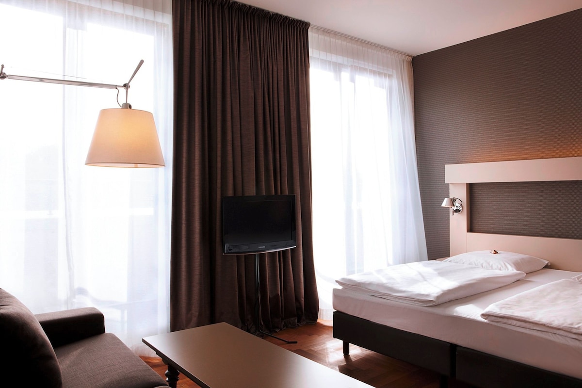 Habitación de hotel con cama, televisor montado en la pared, escritorio y ventana con cortinas.