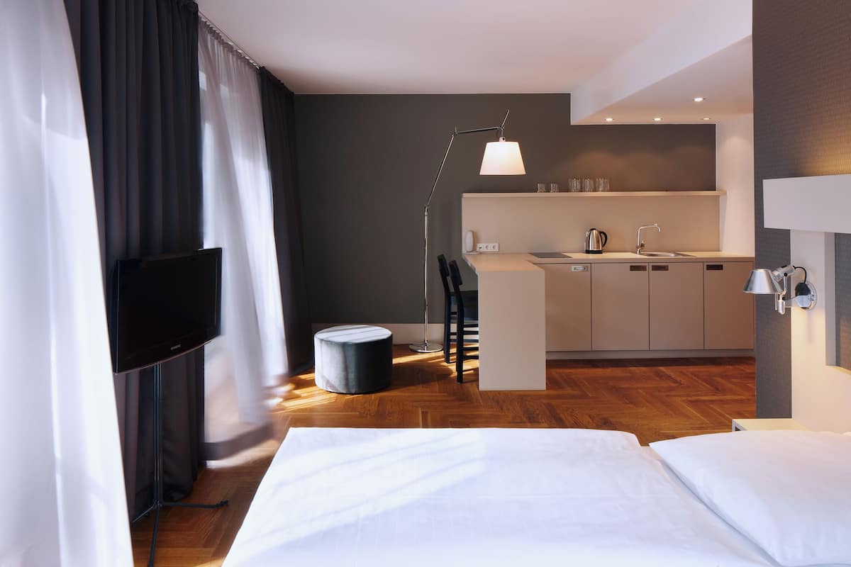 Una habitación de hotel moderna con cama, televisión, escritorio con lámpara, kitchenette y un pequeño taburete redondo.