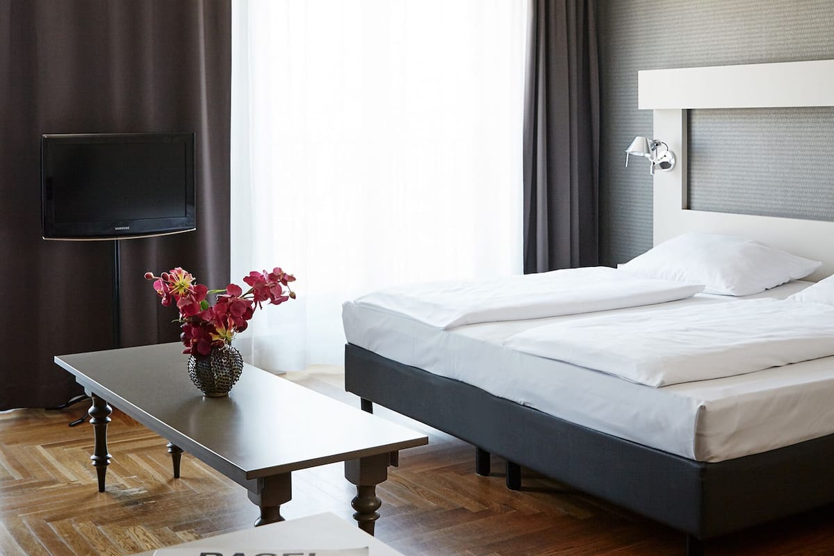 Una habitación de hotel con cama, televisión, una mesa con un jarrón de flores y una revista sobre la mesa.