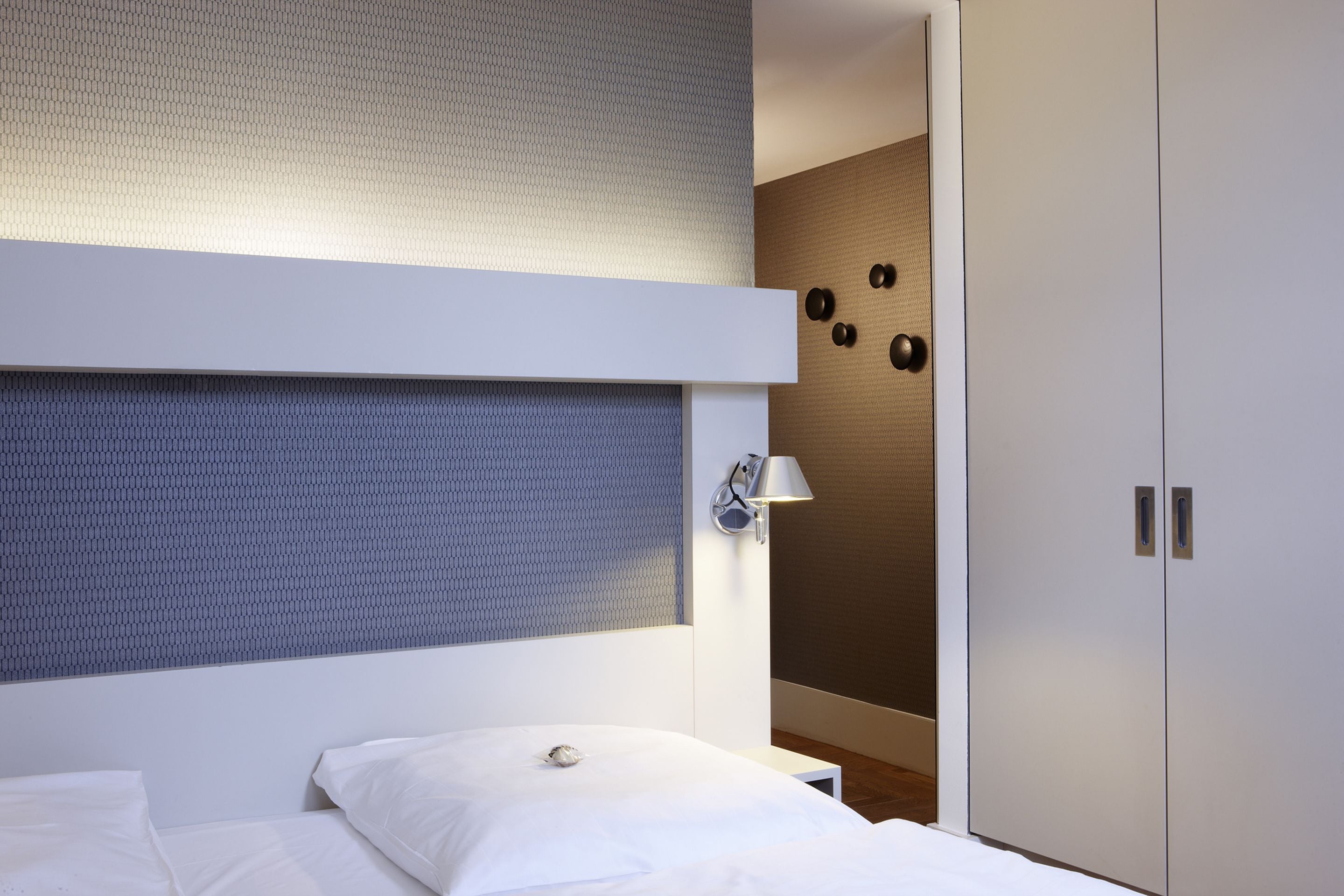 A modern hotel room with a bunk bed, a bedside table, a wall-mounted lamp, and a wardrobe with a mirror.