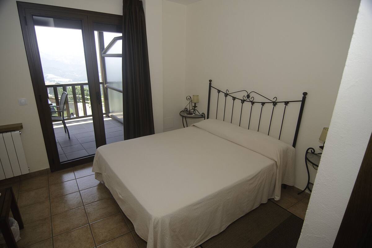 Een slaapkamer met een bed, een nachtkastje, een raam met gordijnen en uitzicht op een balkon.