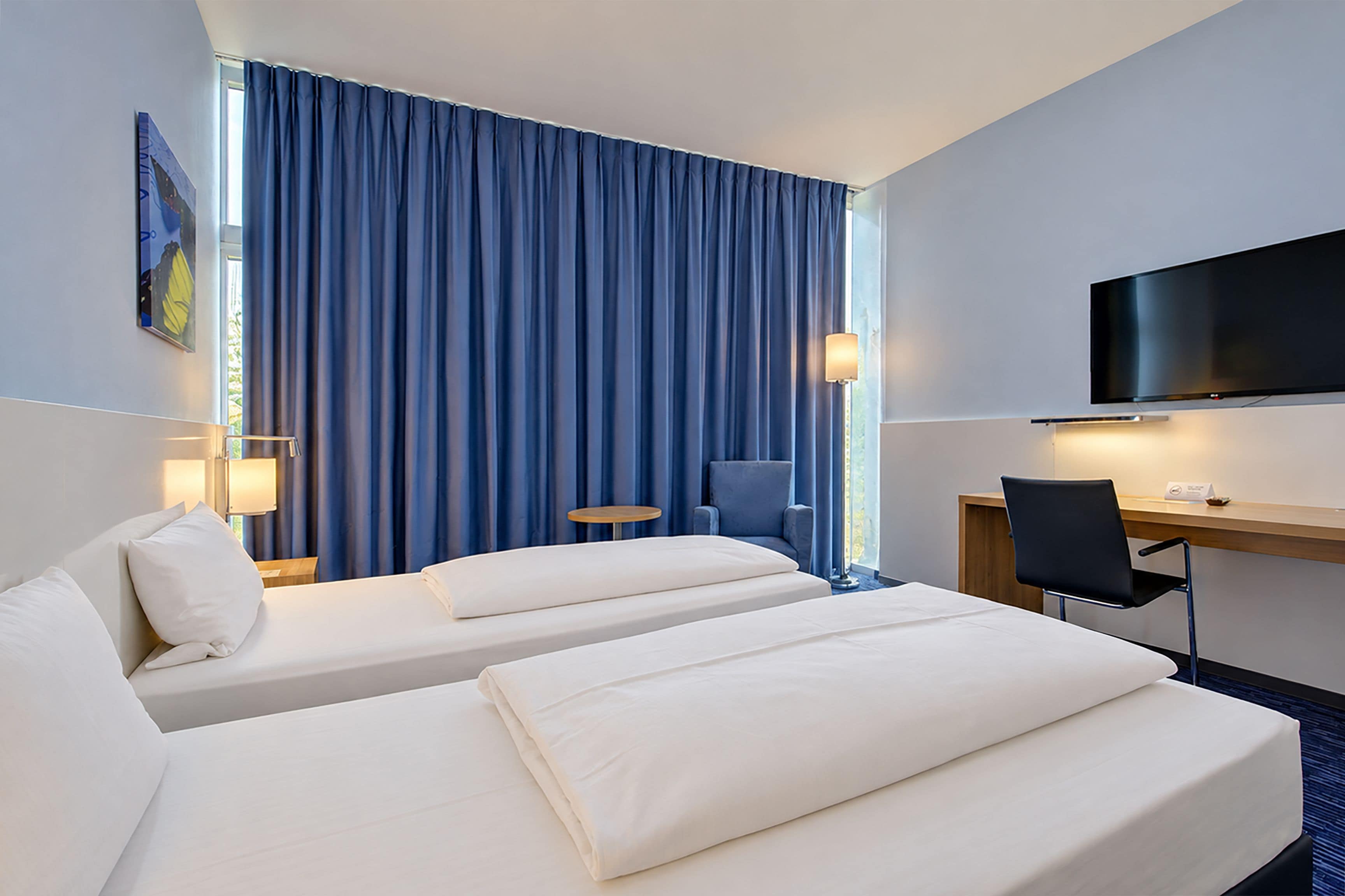 Habitación de hotel con dos camas, un escritorio, una silla, un televisor y una ventana con cortinas.