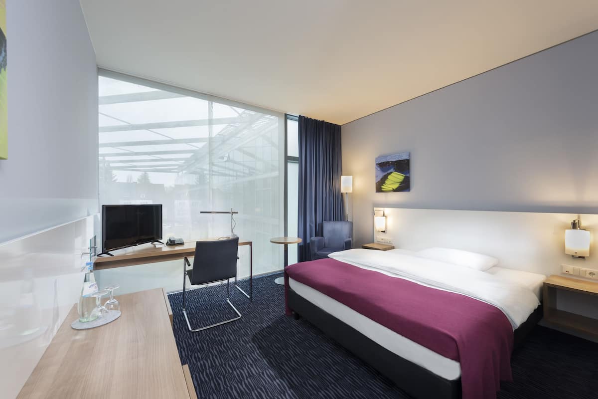 A modern hotel room with a large bed, a desk with a computer, a chair, and a TV.