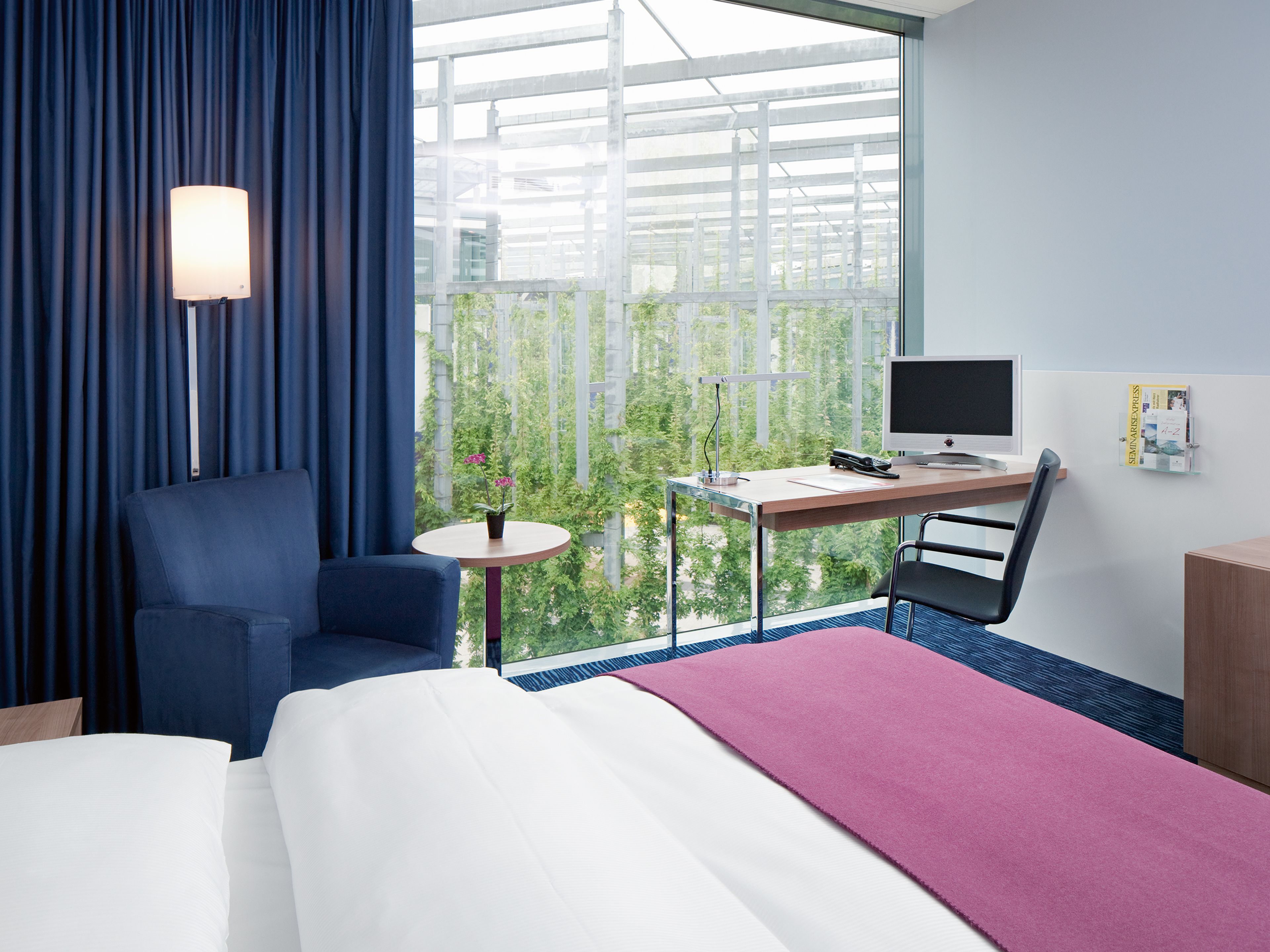 Ein Hotelzimmer mit einem Bett, einem Schreibtisch mit Computer, einem Stuhl, einem kleinen Tisch mit Pflanze und einem Fenster mit Blick ins Grüne.
