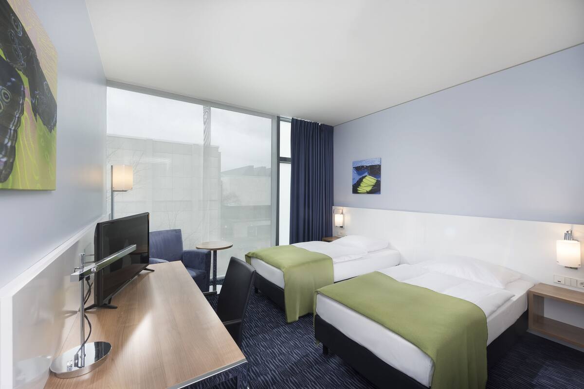 A hotel room with two beds, a desk, a chair, a TV, and a window with curtains.