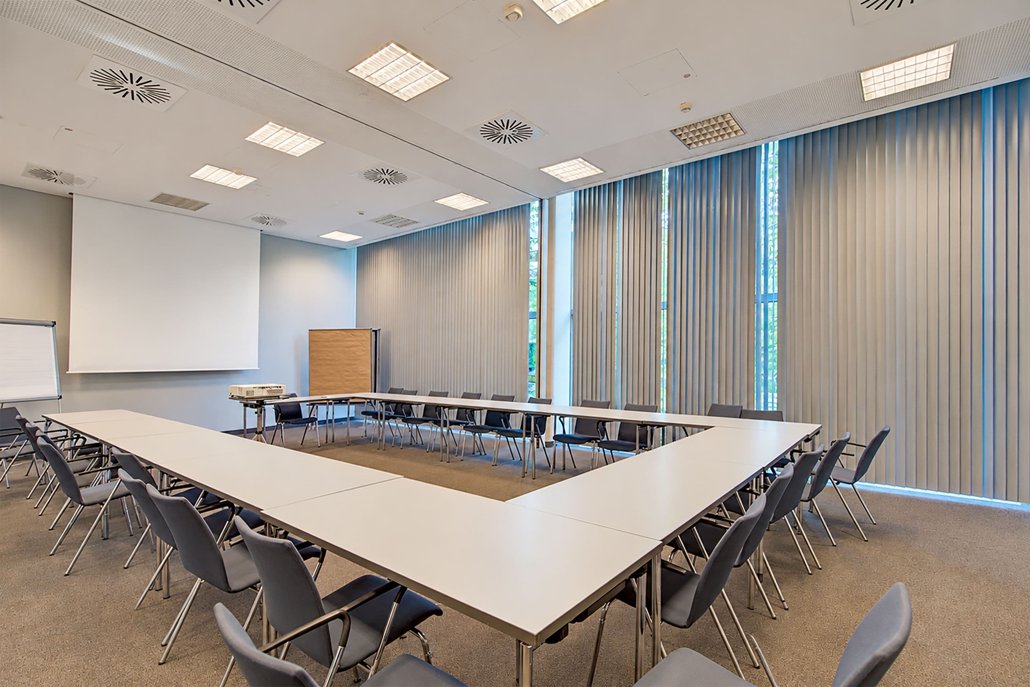 Ein Konferenzraum mit einem langen Tisch, Stühlen, einer Projektionsleinwand und großen Fenstern mit Jalousien.