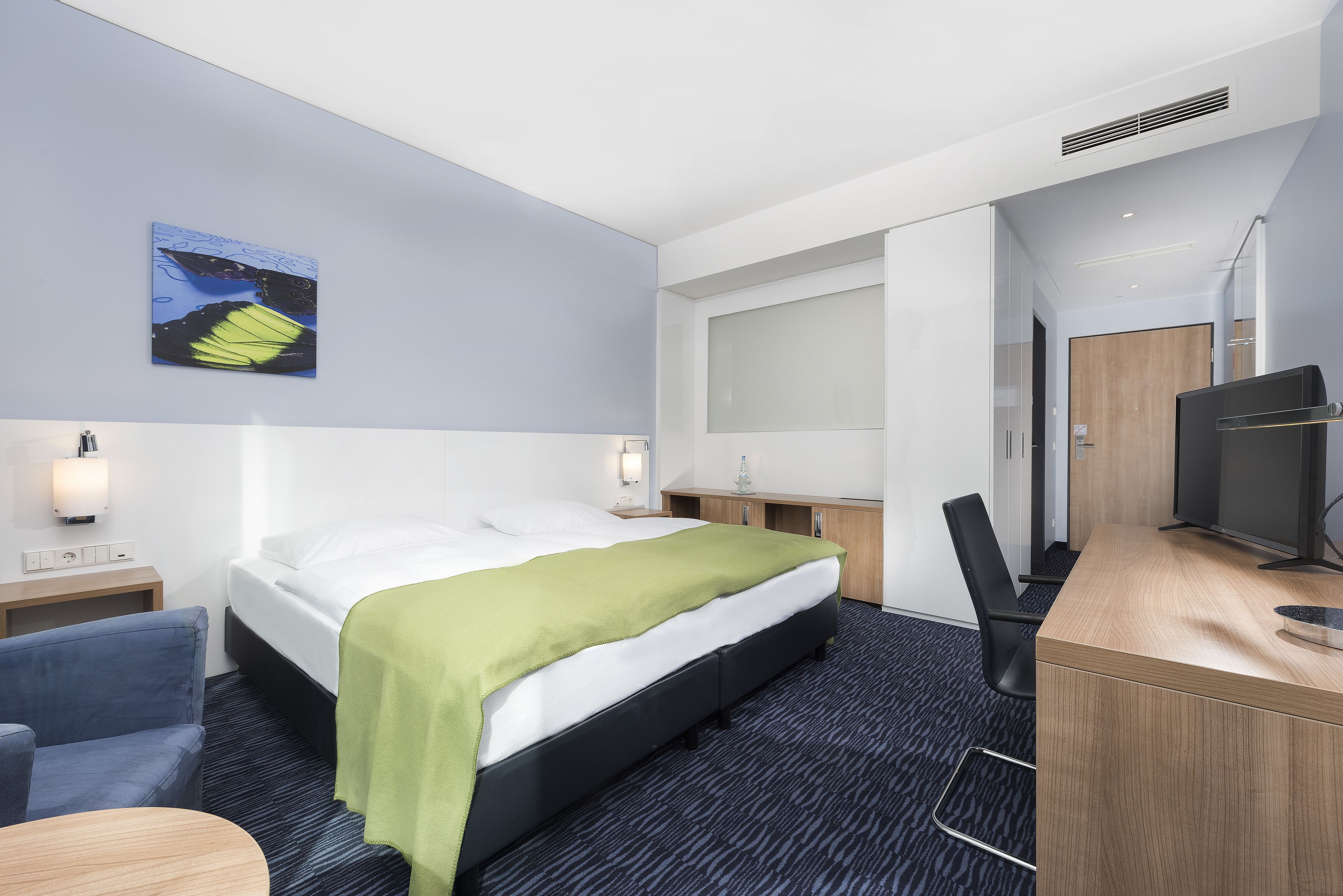 A hotel room with a bed, a desk, a chair, a TV, and a painting on the wall.