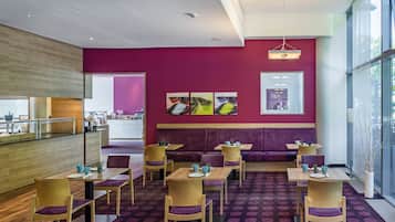 Un espace de restauration moderne avec des murs violets, des tables en bois et des chaises assorties. Des œuvres d’art ornent les murs, et une grande fenêtre laisse entrer la lumière naturelle.