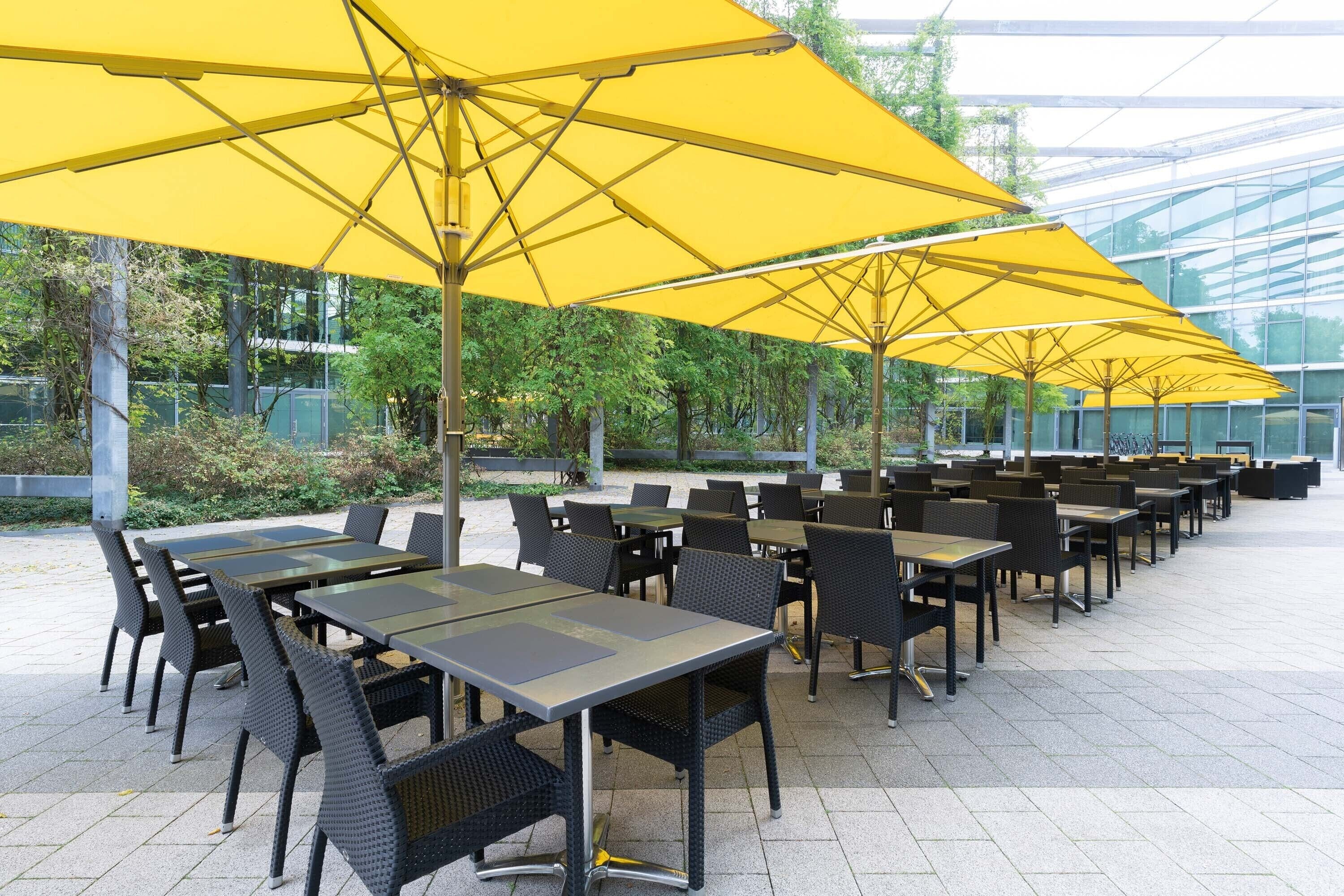 An outdoor seating area with tables and chairs under yellow umbrellas, surrounded by greenery and a glass structure.