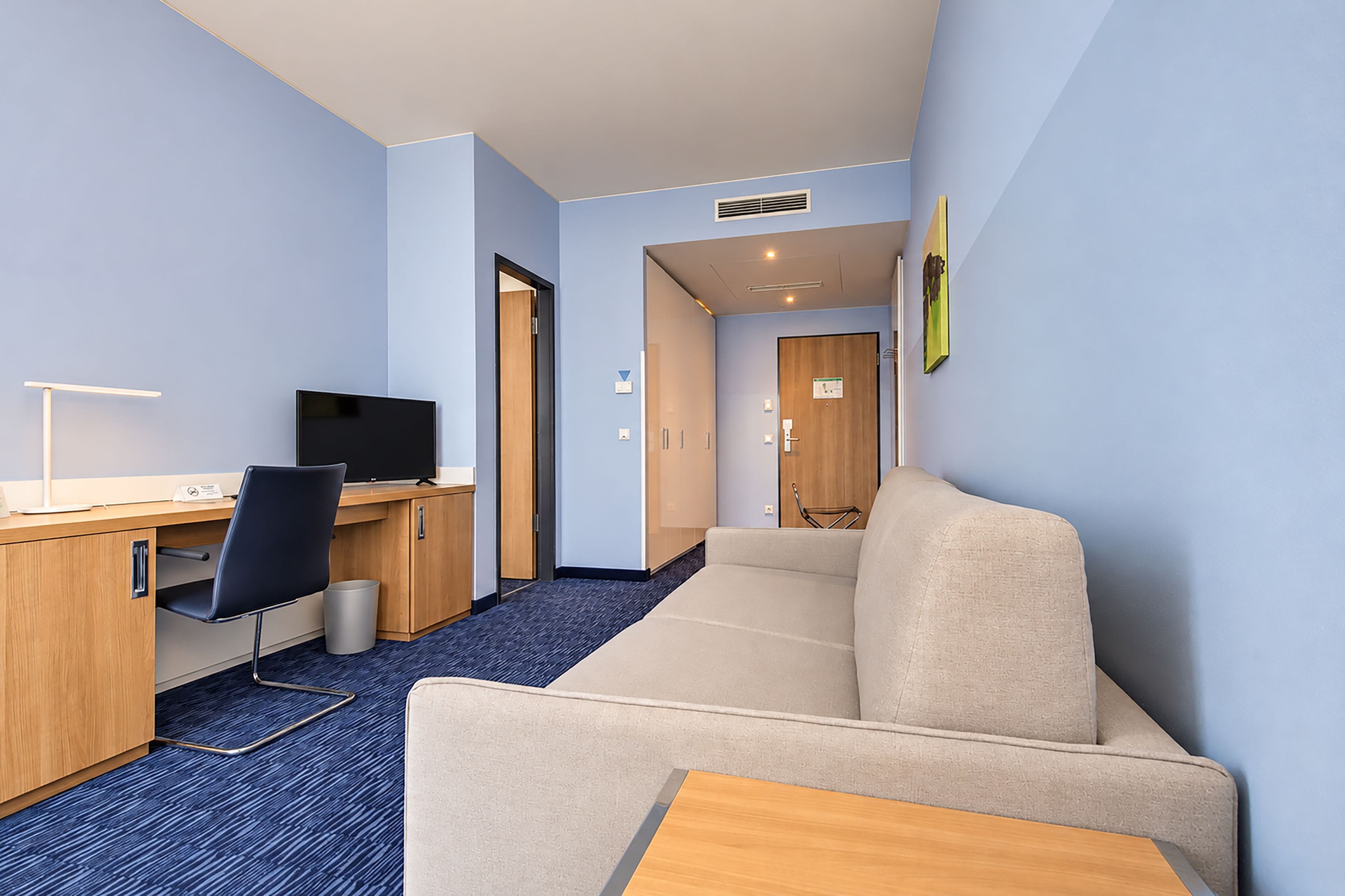 Hotellihuone, jossa on työpöytä, tuoli, televisio, sohva ja pieni pöytä.