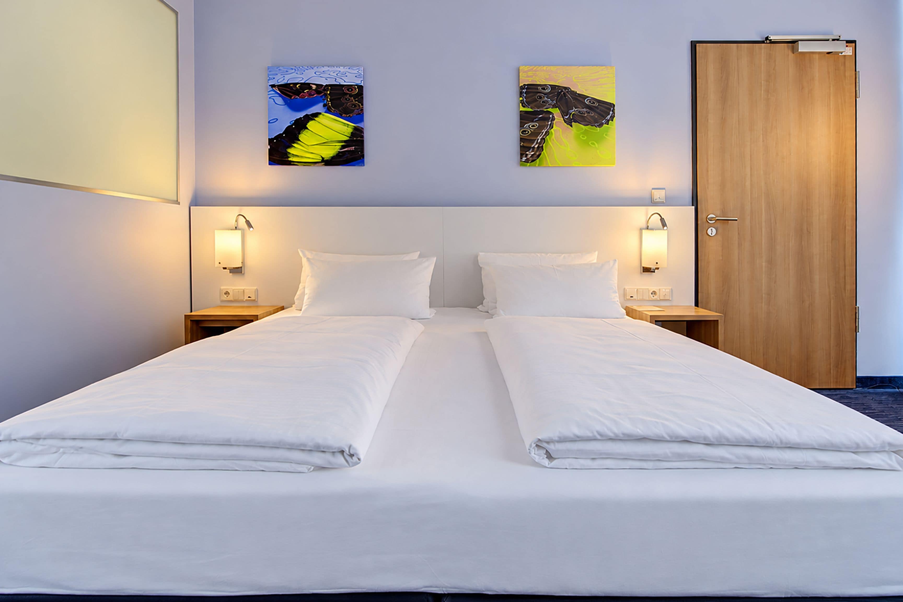 Una cama doble con sábanas blancas, dos mesitas de noche con lámparas y una puerta de madera.