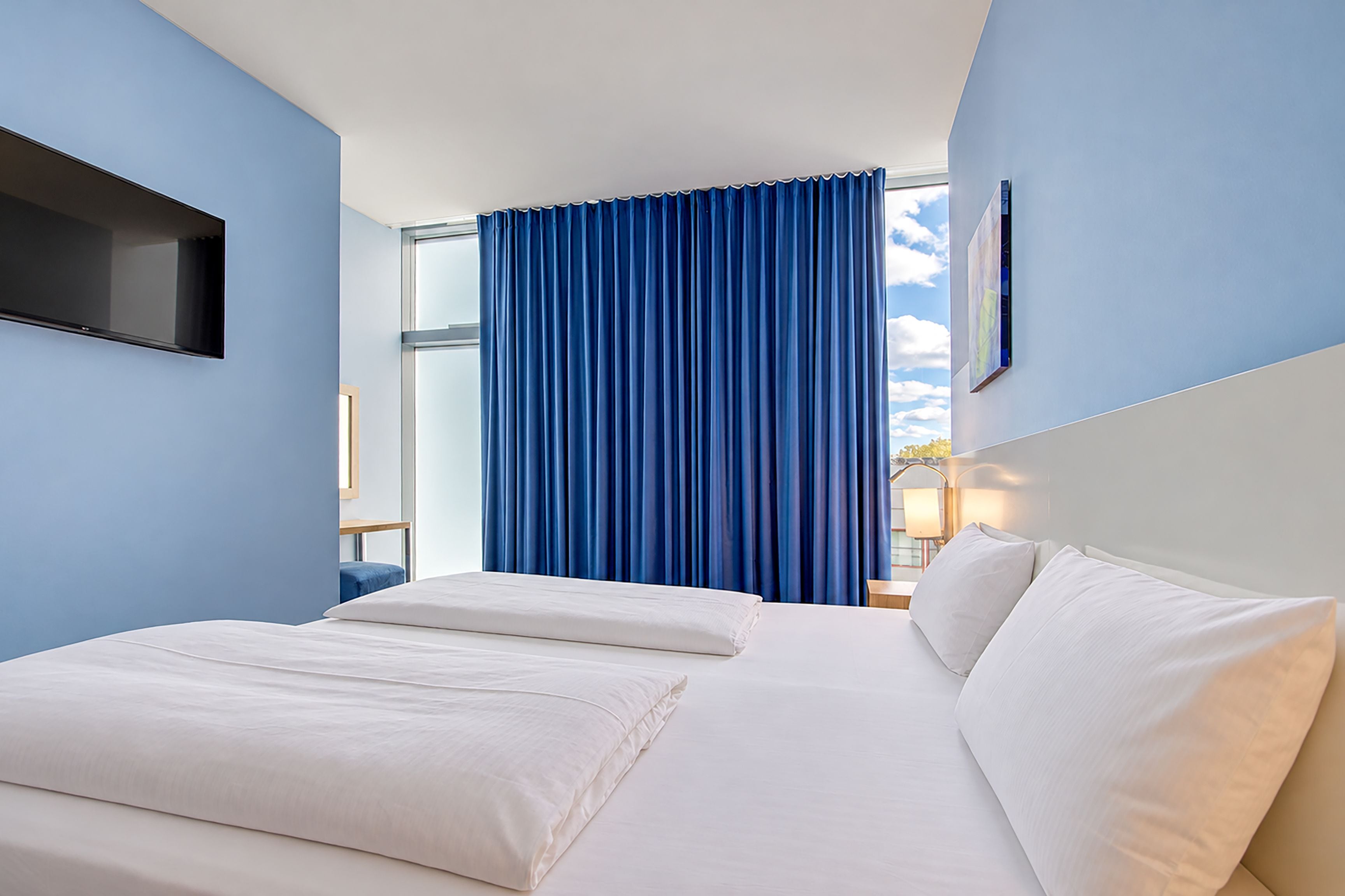 Habitación de hotel con cama, televisor montado en la pared y ventana con cortinas azules.