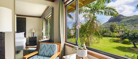 A hotel room with a large window, a bed, a chair, a small table, and a potted plant.