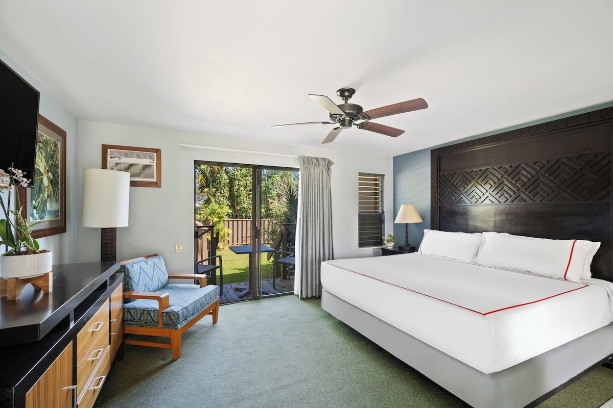 A hotel room with a large bed, a ceiling fan, a desk with a lamp, a chair, and a view of the outdoors.