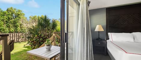 Habitación de hotel con balcón, cama con ropa blanca, mesita de noche con lámpara y vistas a un jardín.