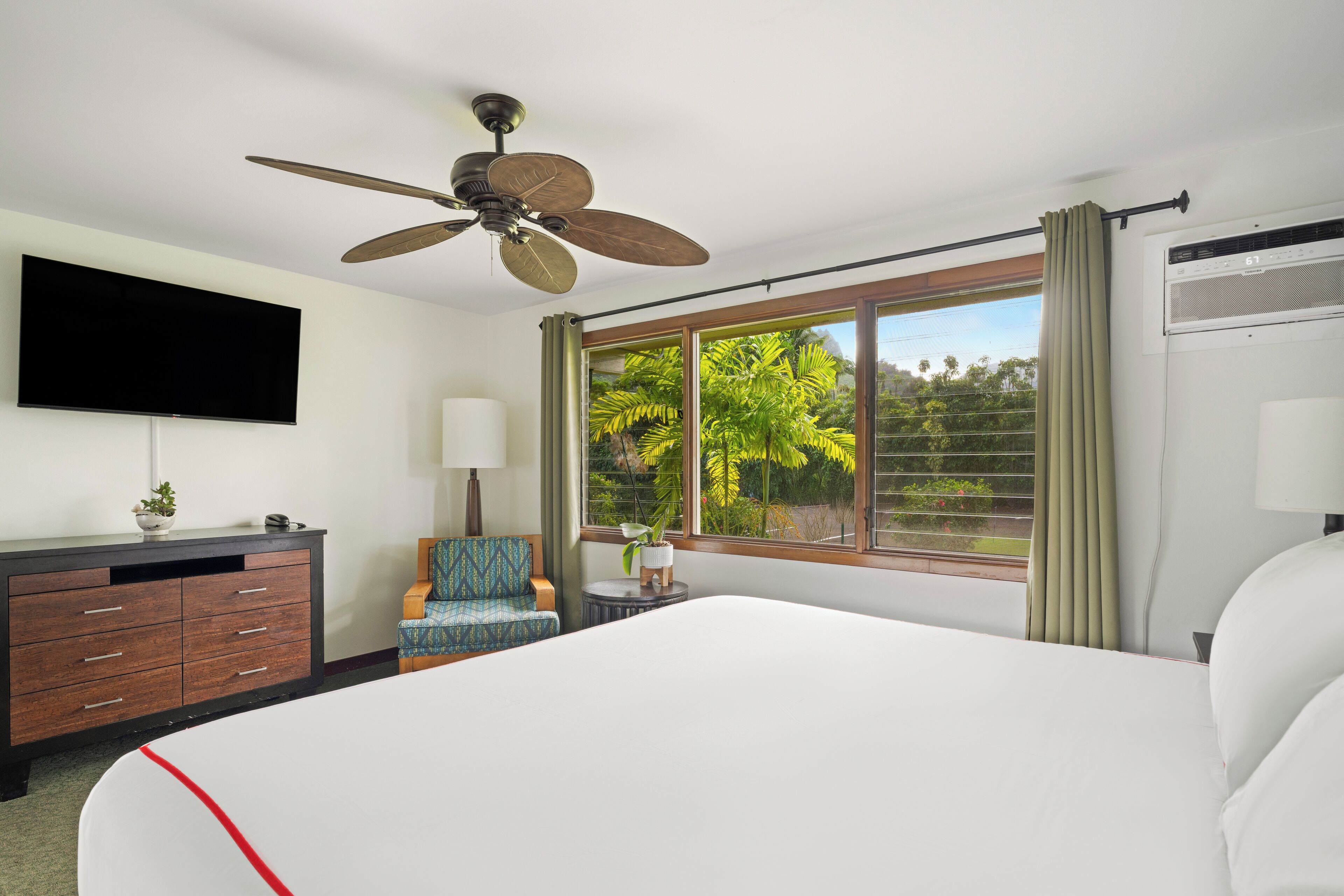 Quarto de hotel com uma cama grande, um ventilador de teto, uma TV de tela plana, uma escrivaninha com luminária, uma cadeira e uma janela com cortinas.