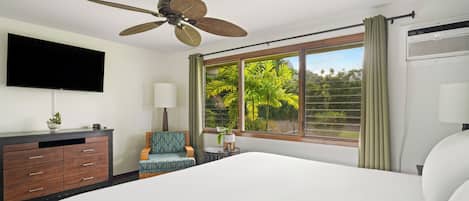 Quarto de hotel com uma cama grande, um ventilador de teto, uma TV de tela plana, uma escrivaninha com luminária, uma cadeira e uma janela com cortinas.
