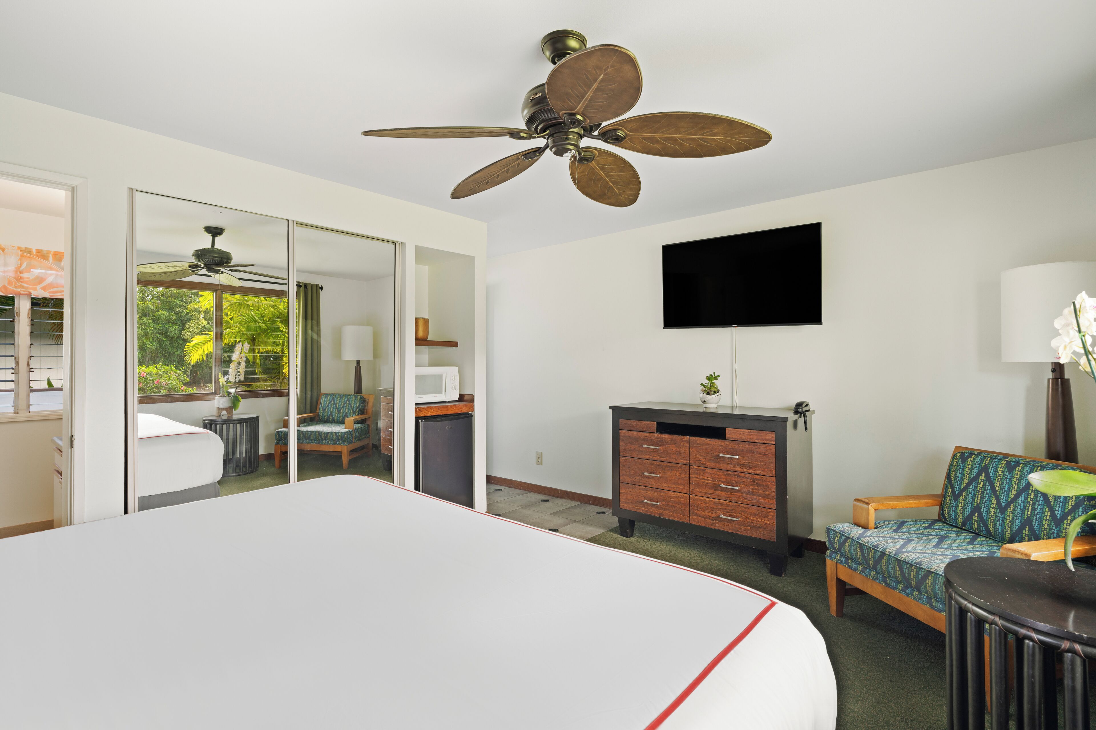 A hotel room with a large bed, a ceiling fan, a flat-screen TV, a wooden dresser, a sofa, and a view of the outdoors.