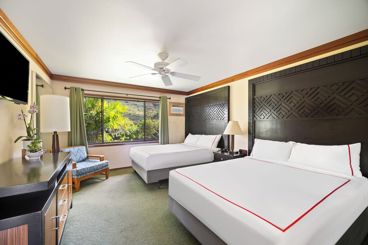 A hotel room with two beds, a desk, a chair, a TV, and a view of greenery outside.