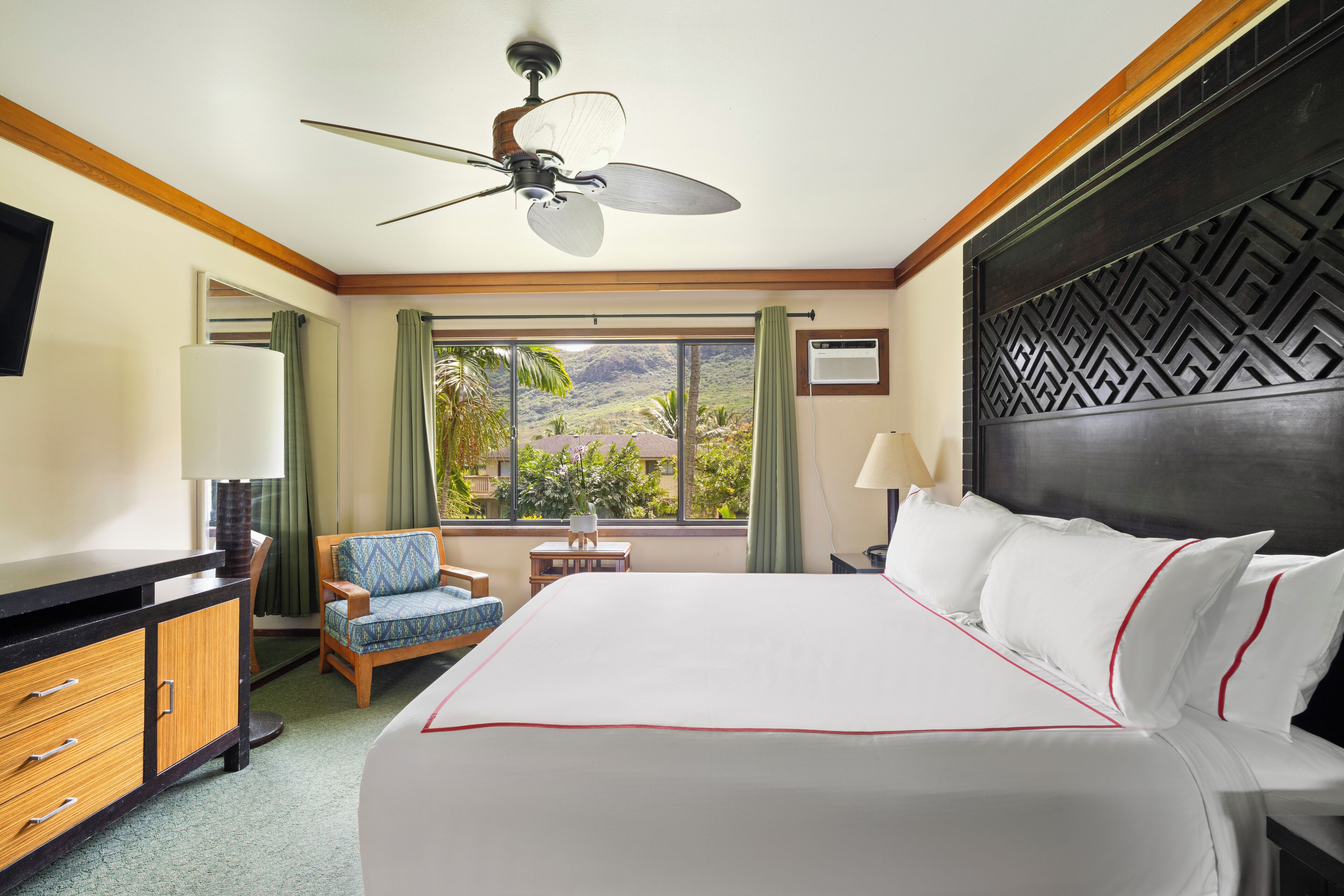 Habitación de hotel con una cama grande, un escritorio con lámpara, una silla, un ventilador de techo y vistas al exterior.
