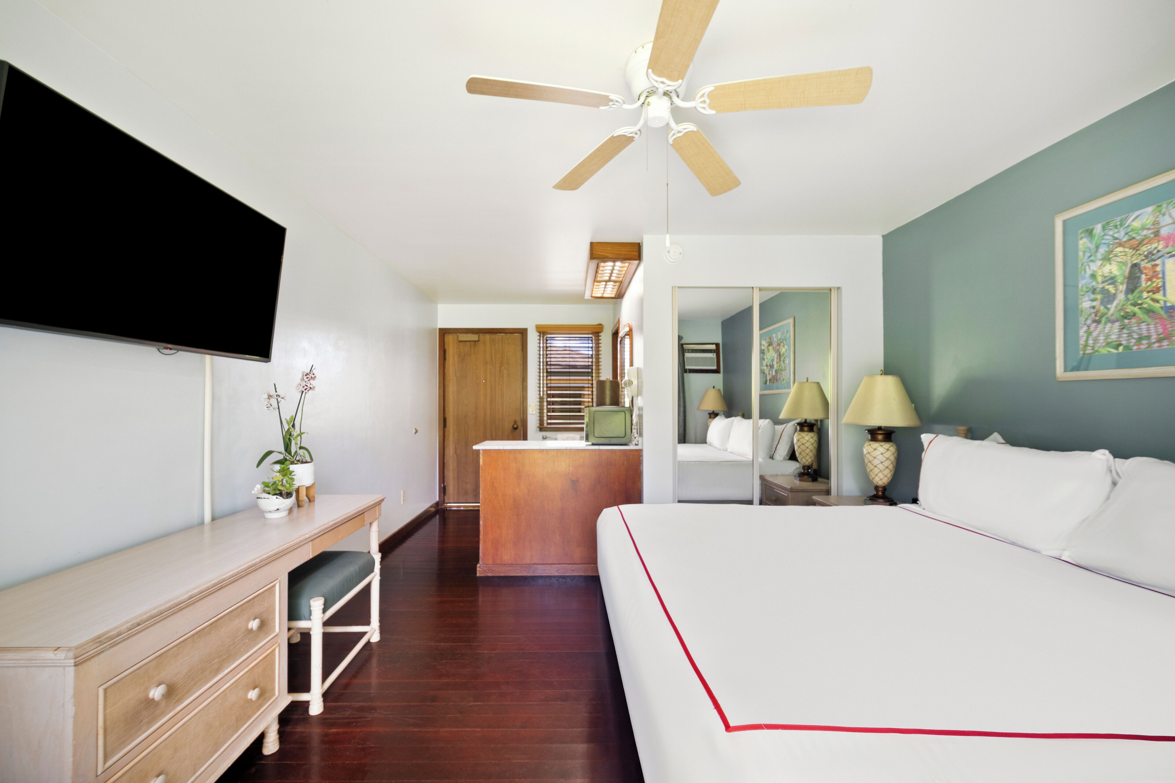 A hotel room with a bed, a desk, a television, a ceiling fan, and a bathroom visible in the background.
