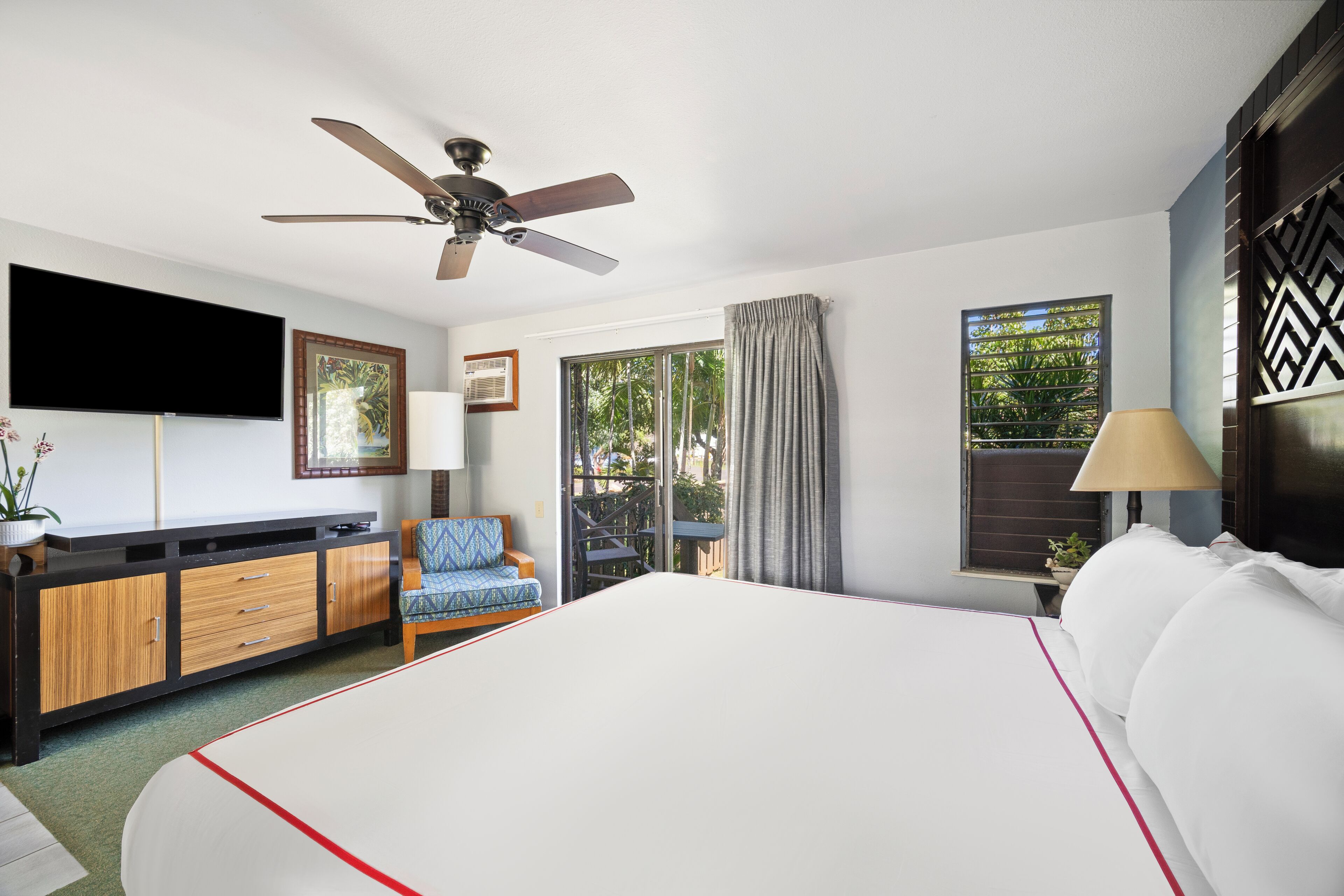 A hotel room with a bed, a ceiling fan, a TV, a desk with drawers, a chair, a window with curtains, and a view of greenery outside.