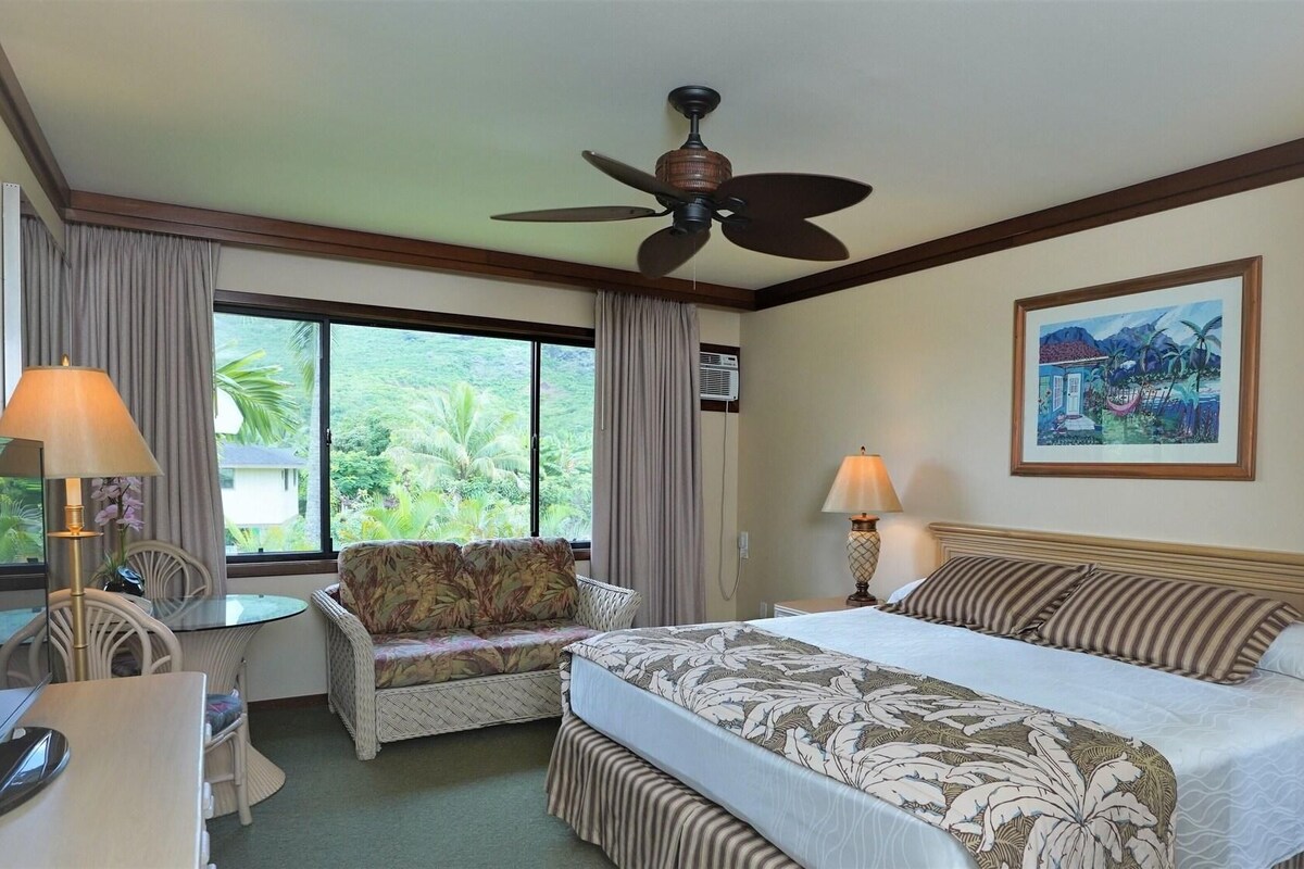 A hotel room with a bed, a desk, a chair, a ceiling fan, and a window with a view of greenery.