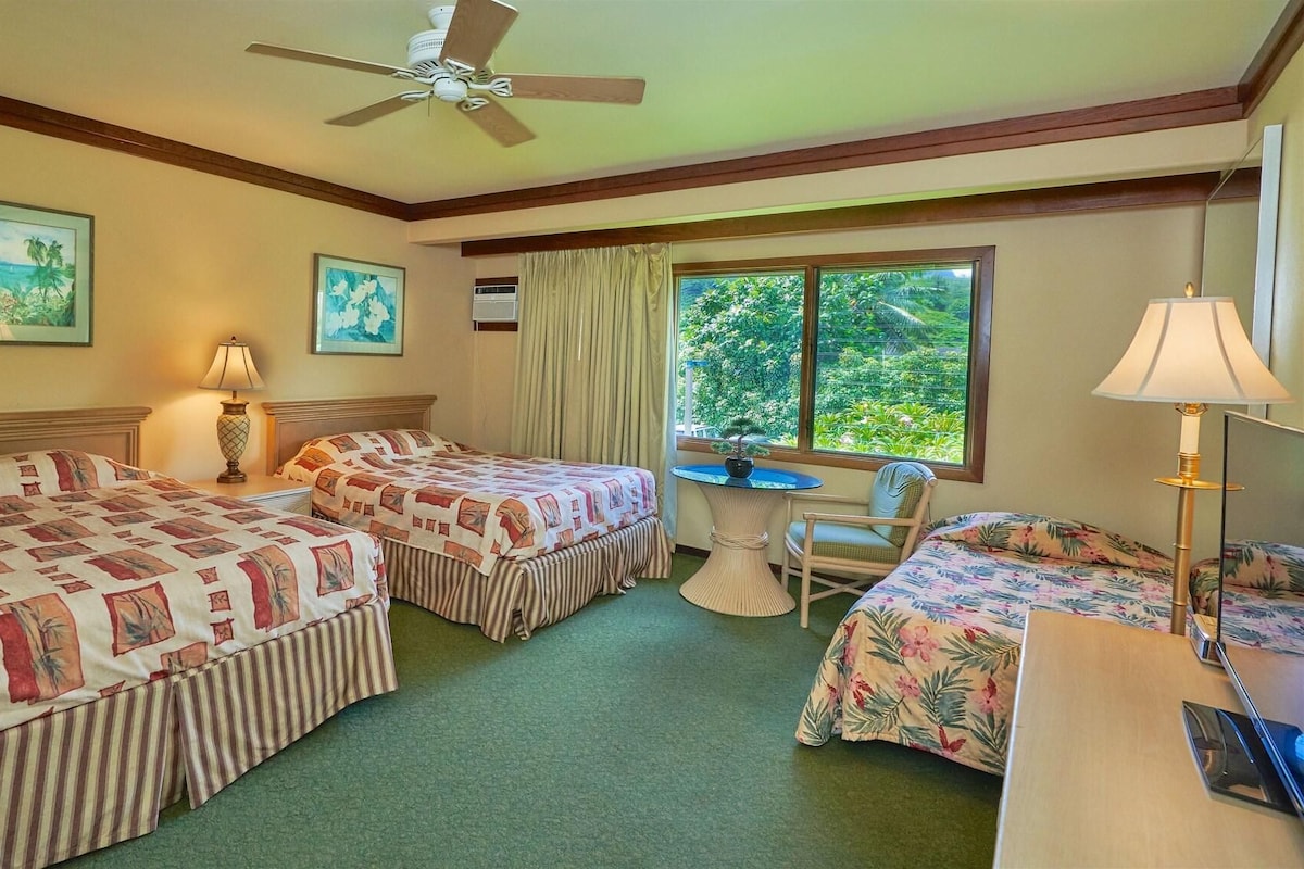 A hotel room with two beds, a small table, a chair, a ceiling fan, and a window with a view of greenery.