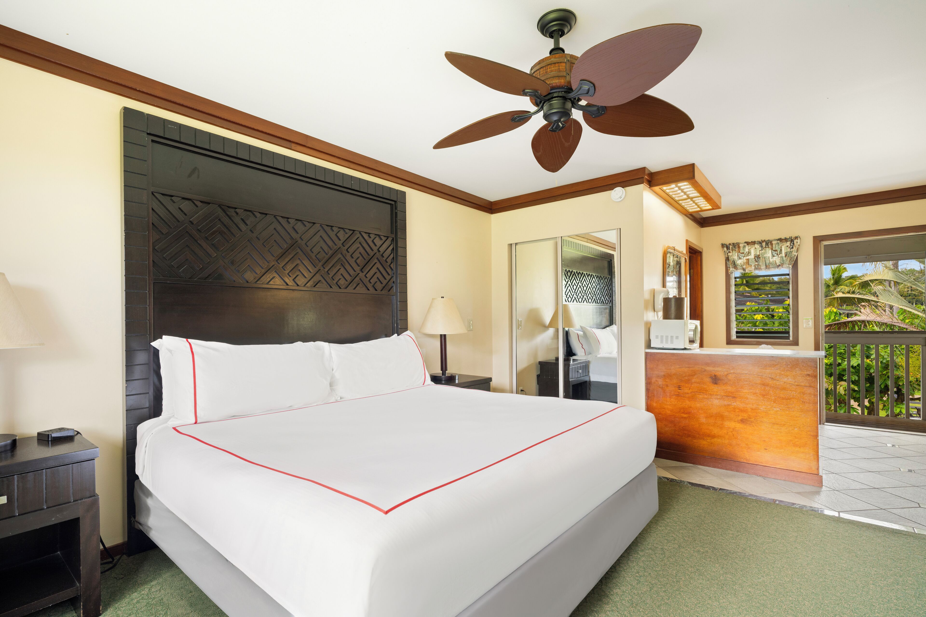 A hotel room with a large bed, a ceiling fan, a wooden headboard, a desk, and a balcony with a view of palm trees.