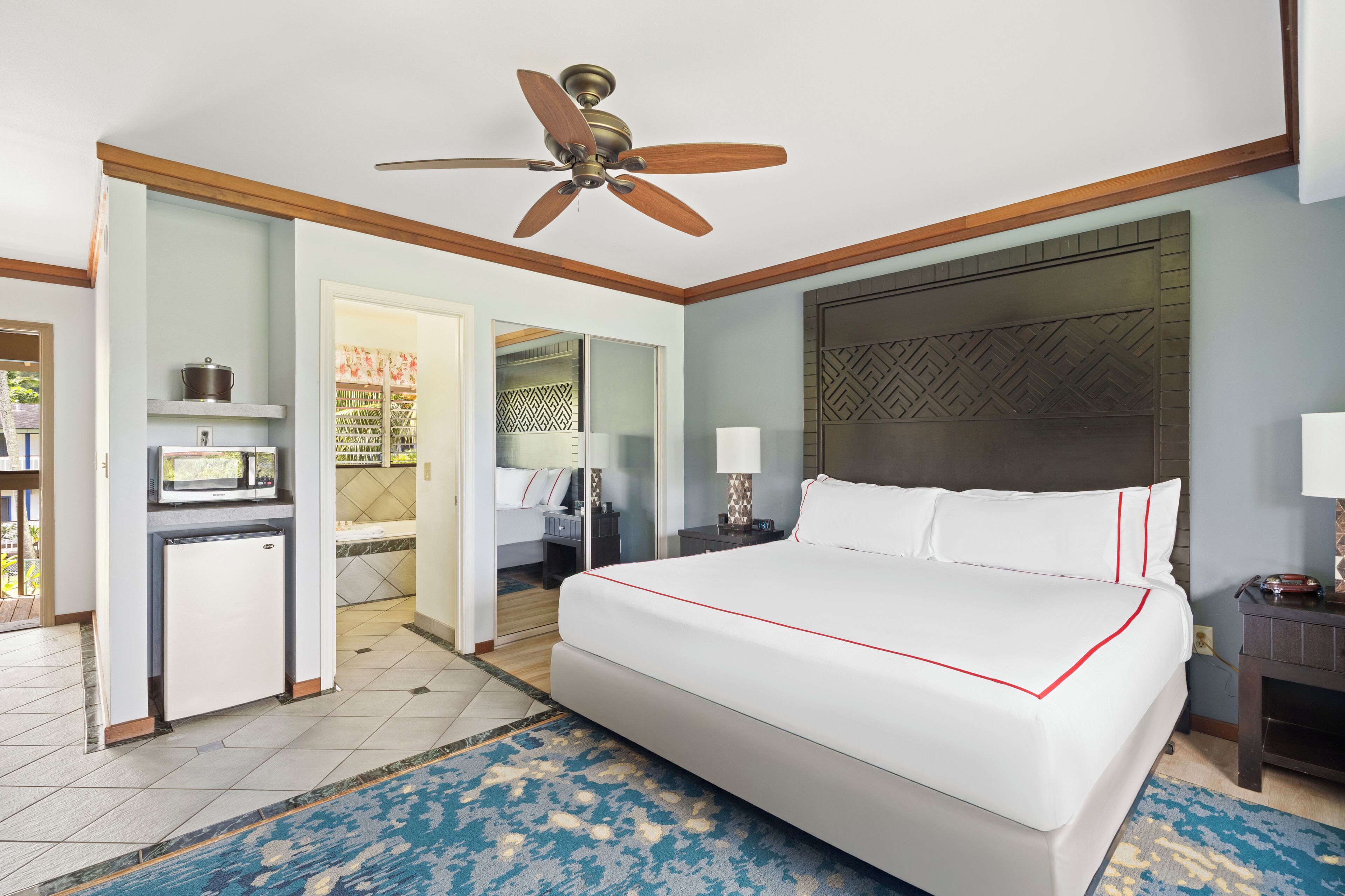 A hotel room with a large bed, a ceiling fan, a small kitchenette, and a bathroom visible through an open door.