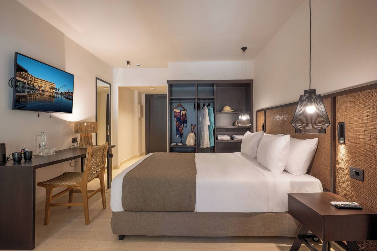 Quarto de hotel moderno com cama, escrivaninha, cadeira, televisão, guarda-roupa e cabideiro.