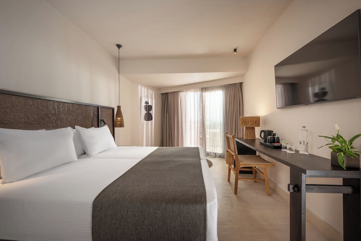 Quarto de hotel com uma cama grande, uma escrivaninha, uma cadeira, uma televisão e uma janela com cortinas.