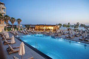 A poolside area with lounge chairs and umbrellas, a building with a pool, and a view of the sea.