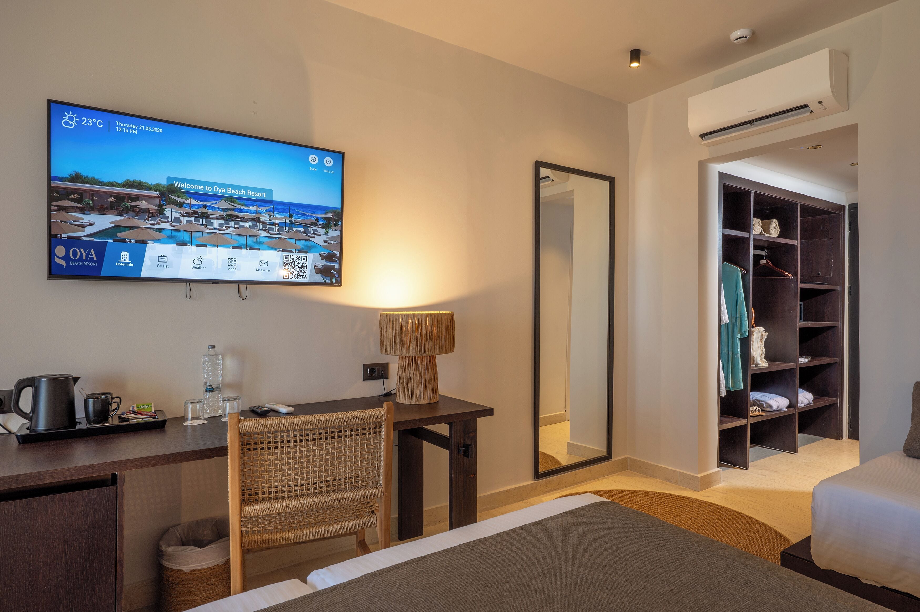 Ein Hotelzimmer mit Fernseher, der einen Bildschirm mit Willkommensgruß des Resorts zeigt, einem Schreibtisch mit Wasserkocher, einem Sessel, einem Nachttisch mit Lampe, einem Kleiderschrank und einem Bett.