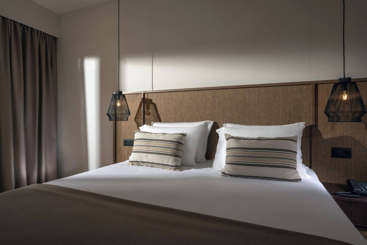 Cama bem arrumada com duas almofadas, mesa de cabeceira com telefone e luminária suspensa.