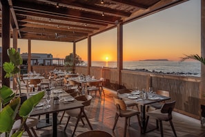 An outdoor restaurant with tables and chairs, a wooden ceiling, and a view of the sunset over the ocean.