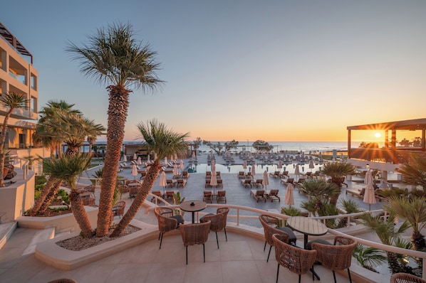 A rooftop terrace with outdoor seating, palm trees, and a view of the sea at sunset.