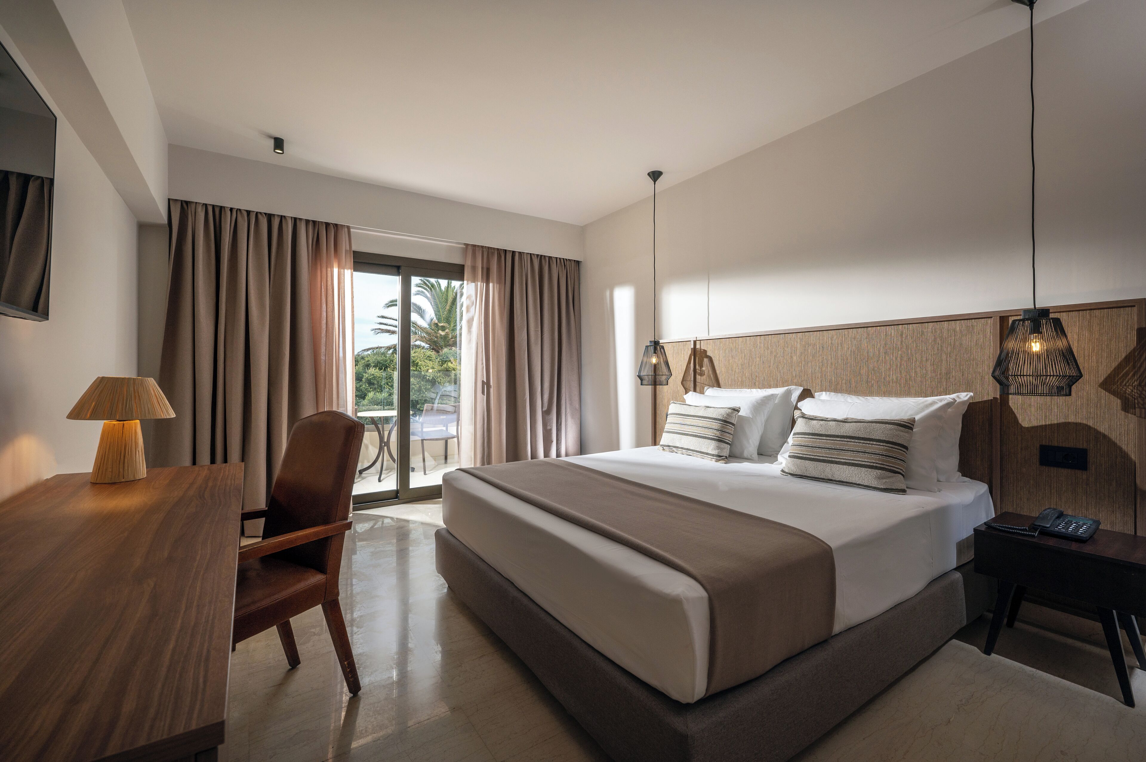 A modern hotel room with a large bed, a desk with a lamp, a chair, and a view of the outdoors through a sliding door.
