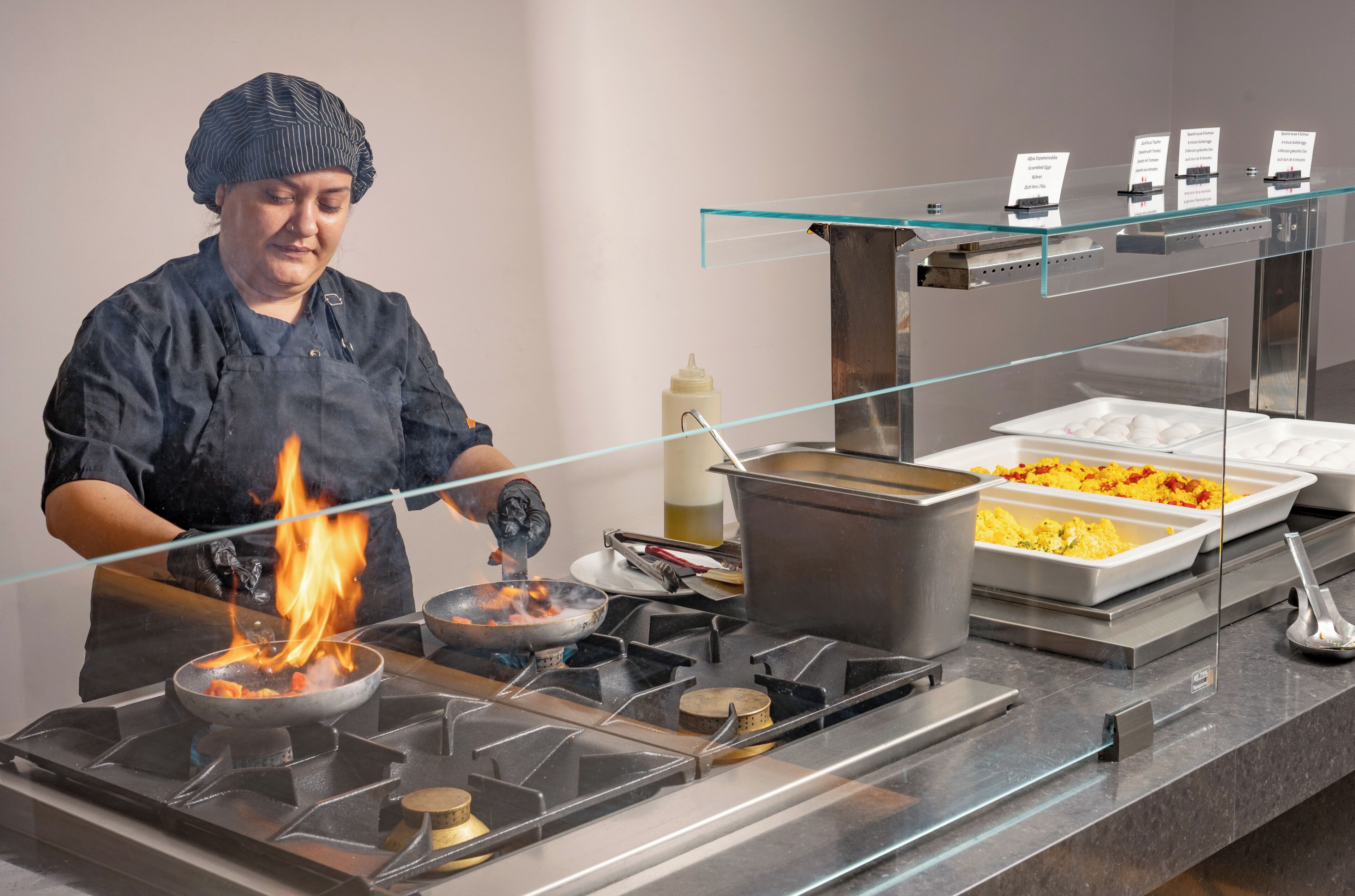 A chef cooking with a pan on a gas stove, a buffet with various dishes, and a glass display case with labels.